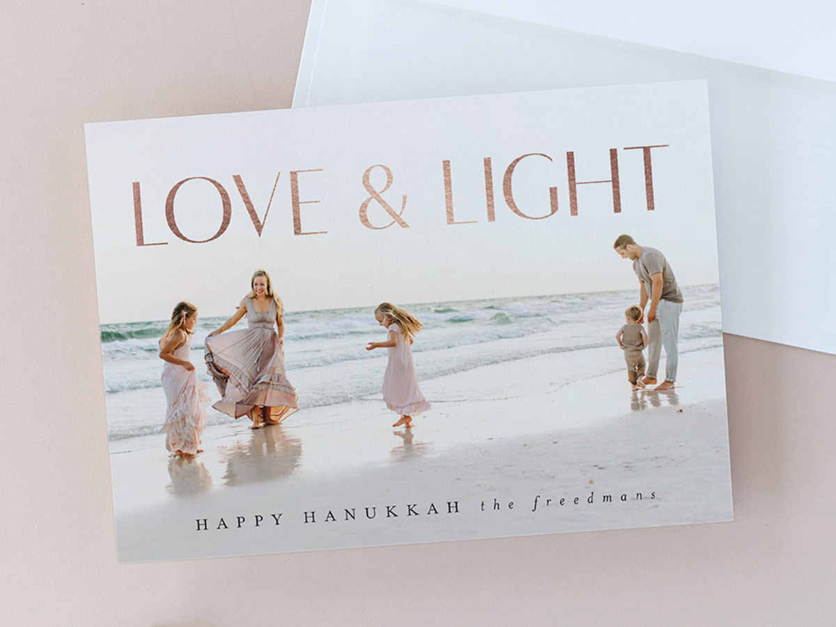 Flat lay image on a dusty pink background with a white envelope and holiday photo card that reads with Love & Light in rose gold foil, Happy Hanukkah, The Freedmans, featuring a young family of five dancing on a sandy beach with waves, design by Jen Huppert Design, available exclusively at minted.com.