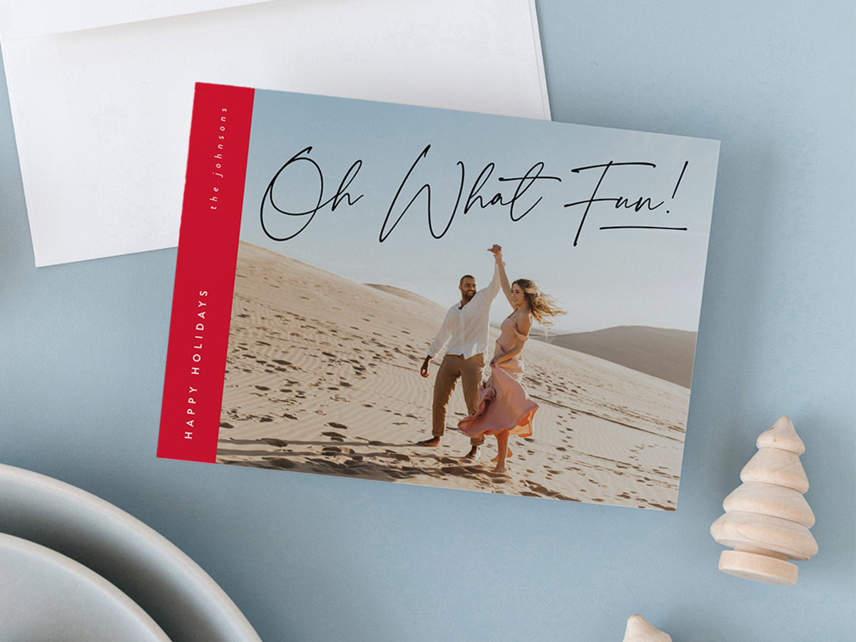 Light blue flat lay image with two white bowls stacked, two small light stained wooden pine trees, white envelope with a holiday photo card that reads Oh What Fun!, Happy Holidays, The Johnsons with a young couple dancing in the desert, design by Jen Huppert Design, available exclusively at minted.com.