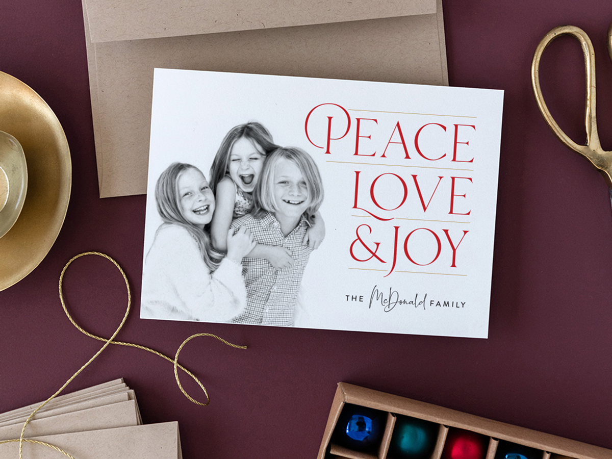 Maroon flat lay with stacked gold bowls, kraft paper envelopes, neutral twine, a box of blue, green, red, and blue round holiday ornaments, with a holiday photo card featuring three children giggling with the words, "Peace, Love, & Joy", The McDonald Family, design by Jen Huppert Design.