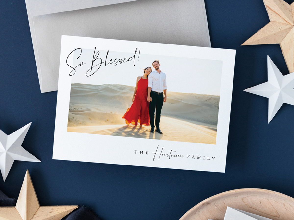 Dark navy blue flat lay background with light wood and white stars, a small wooden bowl, gray envelope, and holiday photo card that reads So Blessed!, The Hartman Family, design by Jen Huppert Design.